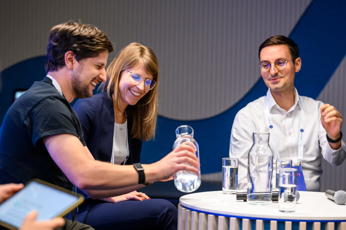 Nahaufnahme eines Gesprächs am Panel-Tisch. Drei Personen sitzen zusammen, auf dem Tisch stehen Wasserkrüge und Gläser.