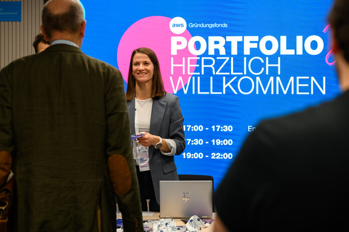 Empfangsbereich mit großem Bildschirm, der „Portfolio herzlich willkommen“ anzeigt. Eine Person steht am Tisch mit Namensschildern und Laptop.