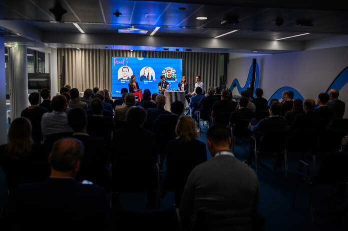Publikum sitzt in einem Konferenzraum und verfolgt eine Panel-Diskussion. Im Hintergrund ist ein großer Bildschirm mit den Namen und Fotos der Panelteilnehmenden sichtbar.