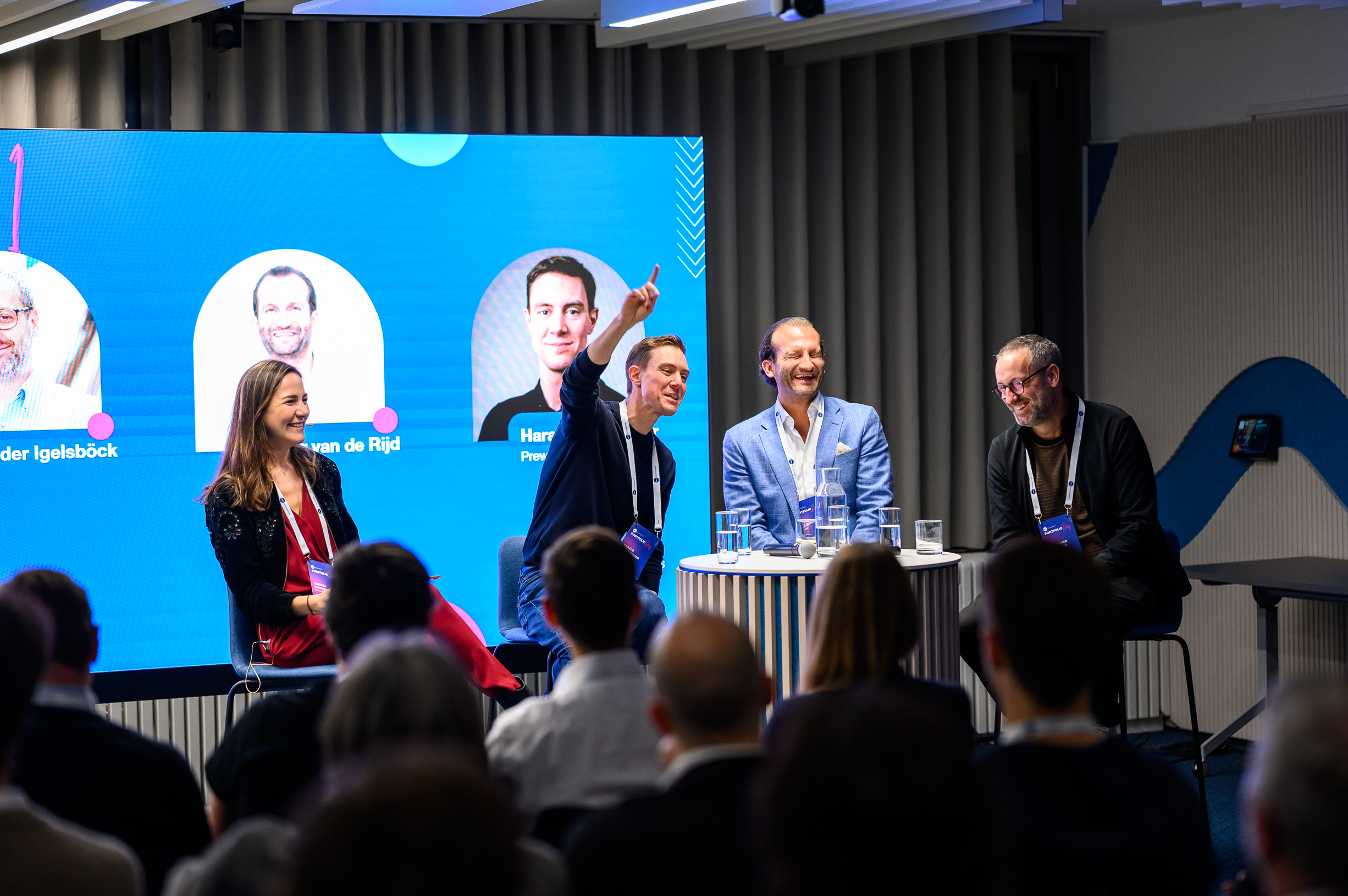 Panel-Diskussion auf der Bühne vor Publikum. Mehrere Sprecher sitzen an einem Tisch, einer zeigt gestikulierend nach oben. Dahinter ist eine Leinwand mit Namen und Fotos.